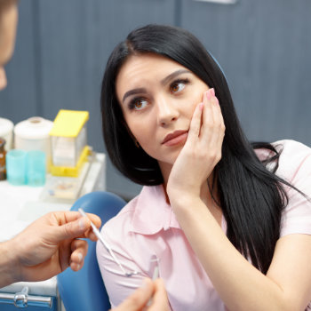 black haired woman with dental pain sitting in a dental chair and touching her cheek