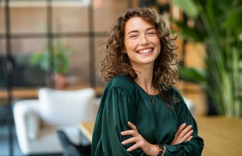 Woman with curly hair wearing a dark green blouse stands indoors with arms crossed, smiling at the camera. Background features plants and a modern, blurred office setting.