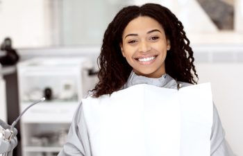 A smiling young woman with dark, curly hair sits in a dental chair, wearing a white paper bib in a dentist's office.